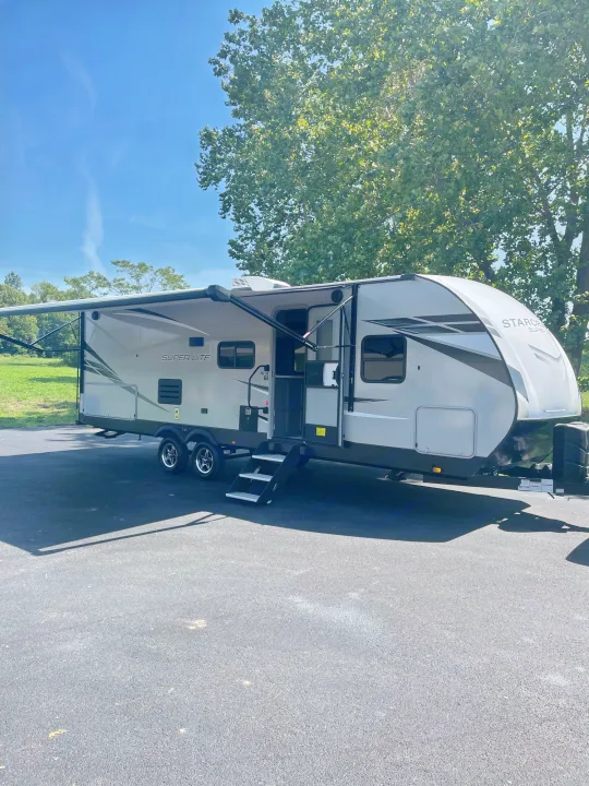 Explore the outdoors! This grey RV rental travel trailer with an extended awning is parked on asphalt under a clear blue sky.. Starcraft Super Lite 2022