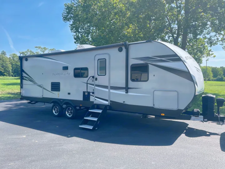 Modern light gray travel trailer RV rental with black steps, parked on asphalt, trees & blue sky background.. Starcraft Super Lite 2022