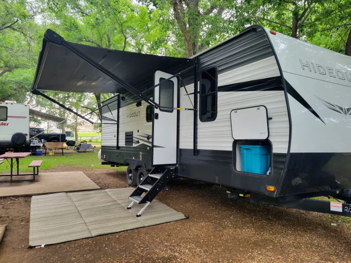 An RV rental is fully set up at a wooded campsite, featuring an extended awning, stairs, and ground mat for outdoor living.. Keystone RV Hideout 2021