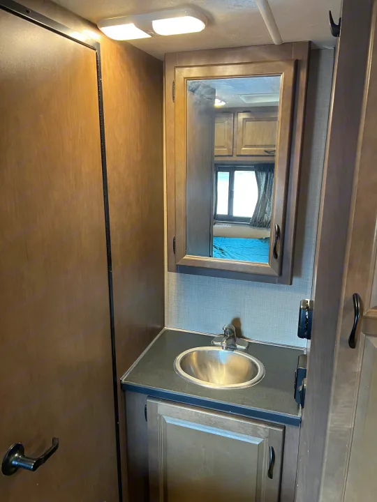 Interior view of an RV rental bathroom vanity, featuring a sink, faucet, mirror, and storage cabinet. A bedroom is visible in the mirror's reflection.. Thor Majestic 2018