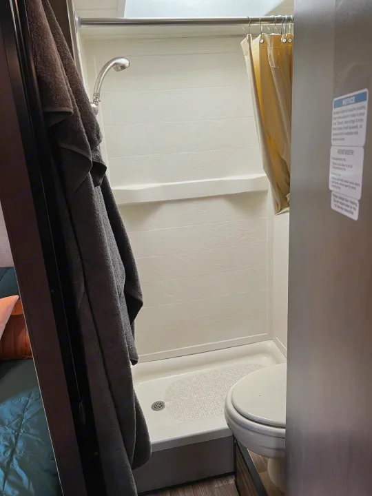 RV rental bathroom interior: compact white shower stall with a curtain, shower head, and toilet visible. A towel hangs nearby.. Thor Majestic 2018