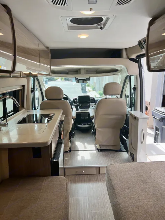 Modern RV rental interior: kitchen with sink/cooktop, swiveling front seats, and rear lounge area.. Thor Sequence 2023