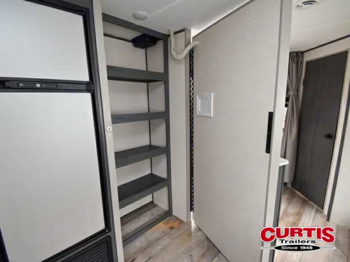 RV rental interior view: compact refrigerator, narrow pantry with dark shelves, and a light door partially open to another room.. Aspen Trail Aspen Trail Trailer 2021