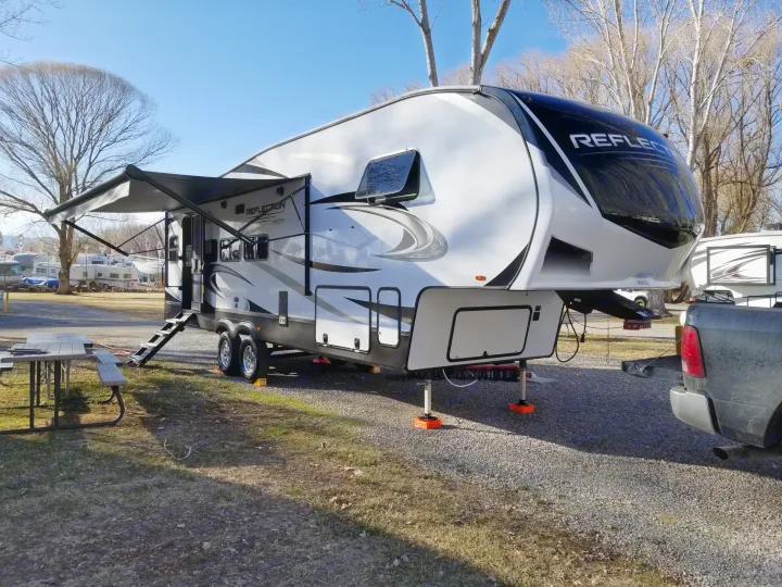 White and black fifth-wheel RV set up at a sunny gravel campsite with an extended awning, ready for your rv rental adventure.. Grand Design Reflection 2021