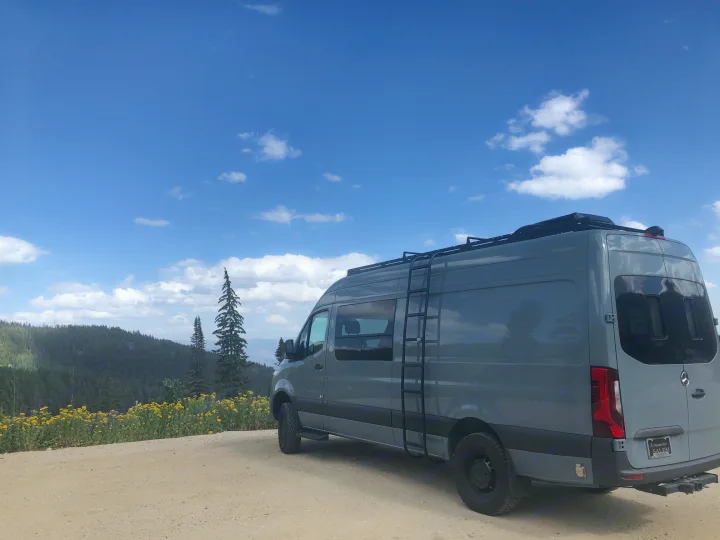 Grey Mercedes Sprinter RV rental on a mountain dirt road, surrounded by wildflowers & pines under blue sky.. Mercedes Sprinter 2022