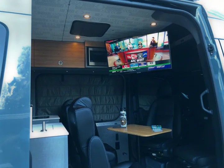 Spacious RV rental interior with a TV displaying AFC West football, kitchenette, and comfortable seating. Perfect for road trips!. Mercedes Sprinter 2022
