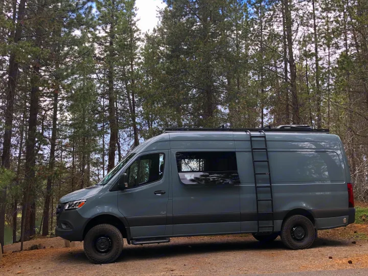 Gray Sprinter RV rental van with off-road tires and side ladder, parked in a scenic pine forest.. Mercedes Sprinter 2022