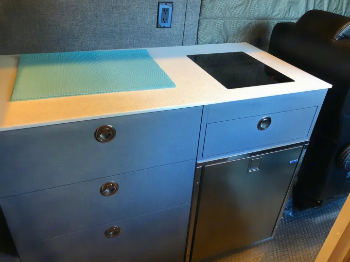 Inside an RV rental: a compact kitchen with induction cooktop, built-in fridge, and textured blue counter mat.. Mercedes Sprinter 2022