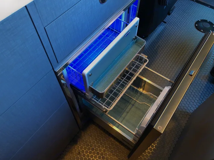 Organized RV rental cooling: a blue-lit Isotherm drawer fridge/freezer with wire baskets opens from textured cabinetry, showing rubber flooring.. Mercedes Sprinter 2022