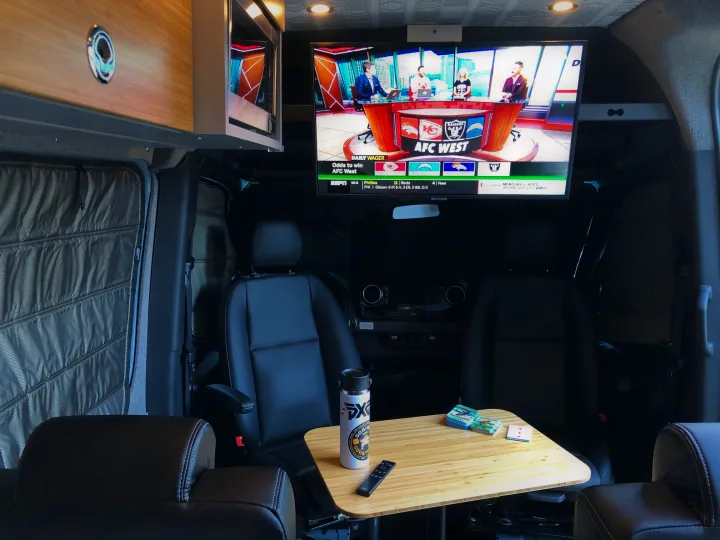Cozy rv rental interior with a big screen TV showing sports, swivel chairs, and a small table. Ideal for travel entertainment.. Mercedes Sprinter 2022