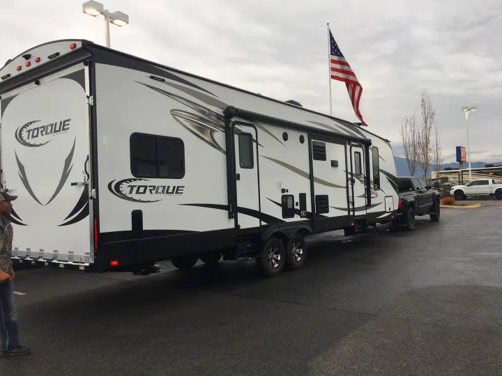 Large white Torque by Heartland travel trailer/toy hauler, perfect for an RV rental, hitched to a pickup truck under cloudy skies.. Heartland Torque 2017
