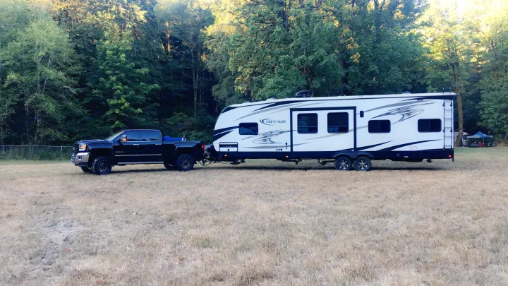 Black pickup towing a white Torque RV in a sunny, grassy field beside lush trees. Ideal rv rental for outdoor travel.. Heartland Torque 2017