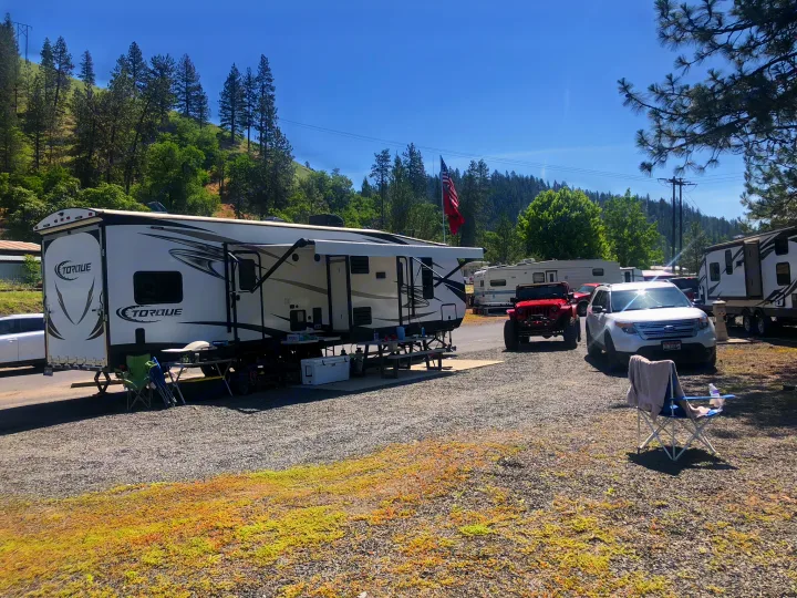 A sunny rv rental campsite shows a large white travel trailer parked on gravel, surrounded by mountains & pine trees.. Heartland Torque 2017