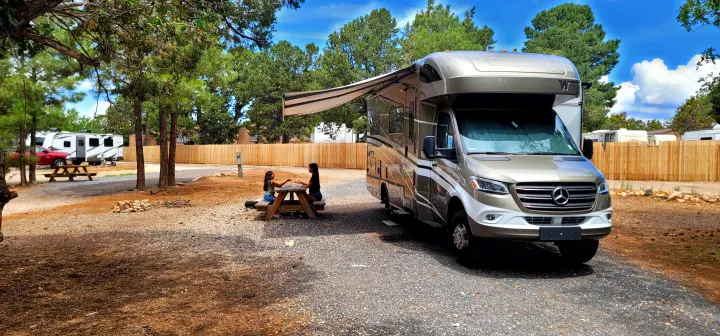 Two people enjoy a picnic at their RV rental campsite, featuring a modern motorhome with an extended awning.. Winnebago Navion 2022