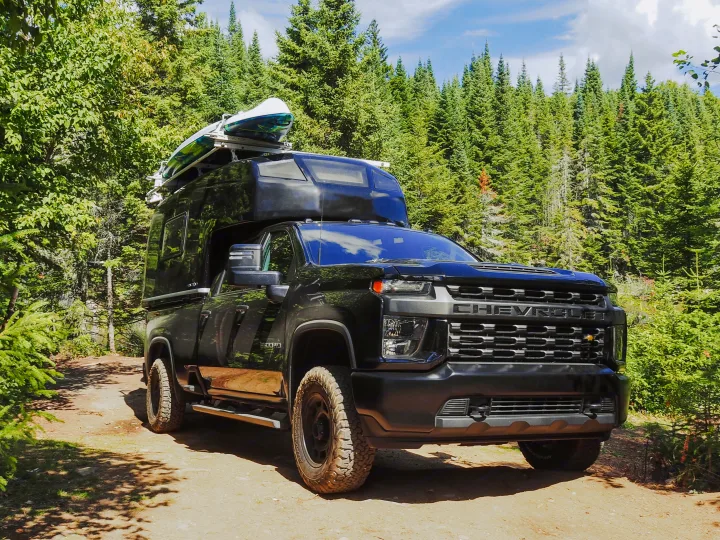 Rugged black truck camper for your next RV rental adventure. Parked off-road in a dense forest with a kayak on top.. Chevrolet silverado 2500 2022