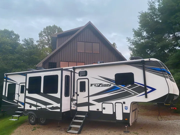 Spacious Fuzion fifth-wheel camper, ideal for an RV rental, parked on gravel with a unique house and trees behind it.. Keystone RV Fuzion Toy Hauler 2021