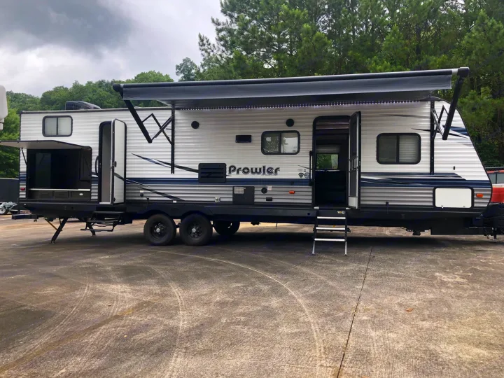 A spacious light grey Prowler travel trailer RV rental with an extended awning and an open outdoor kitchen, parked on asphalt.. Heartland RVs Prowler 2020