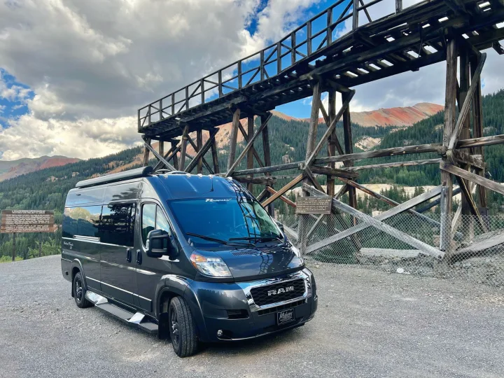 Dark RV rental parked at a historic wooden mining structure, with red-streaked mountains under cloudy skies.. Dodge Other 2020