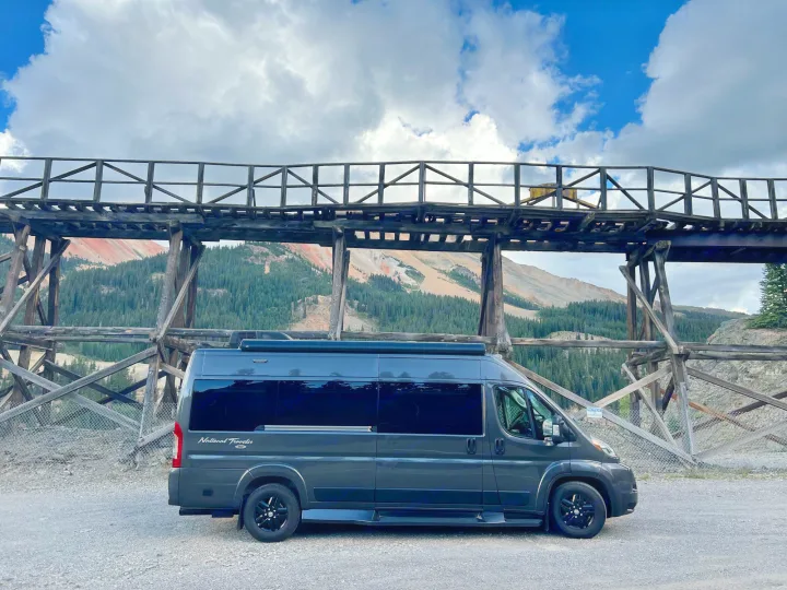 Dark camper van (RV rental) by old wooden trestle, with colorful mountains & cloudy skies. Scenic travel.. Dodge Other 2020