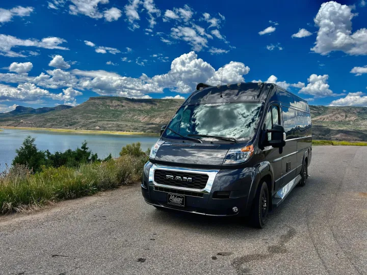 Black RV rental parked by a scenic lake with mountains under a bright blue sky and fluffy clouds.. Dodge Other 2020