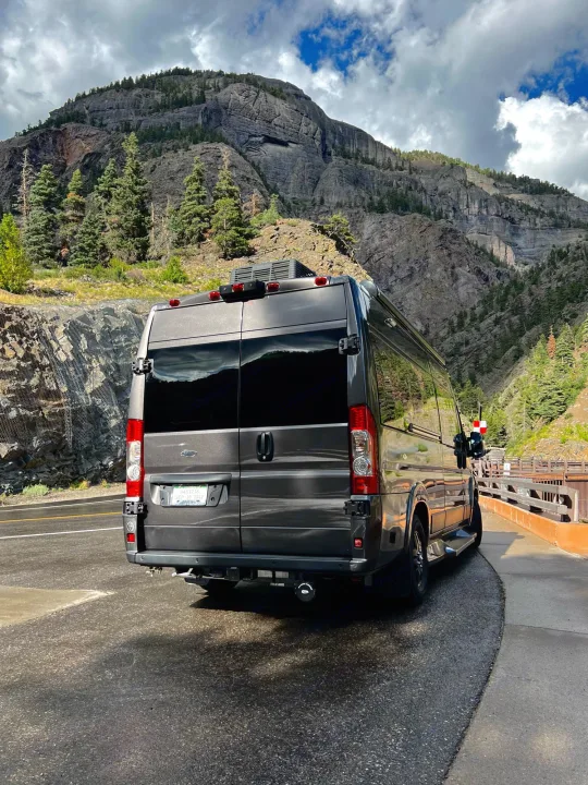 Dark grey RV rental parked on a mountain road, showcasing rocky peaks, pine trees, and cloudy skies.. Dodge Other 2020