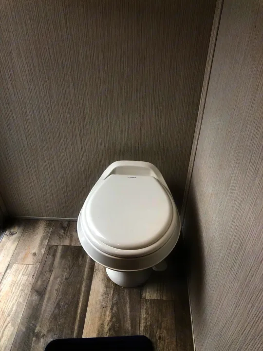 Compact white Dometic toilet in an RV rental bathroom, featuring light grey wood-grain walls and matching floor.. Heartland RVs Prowler 2020