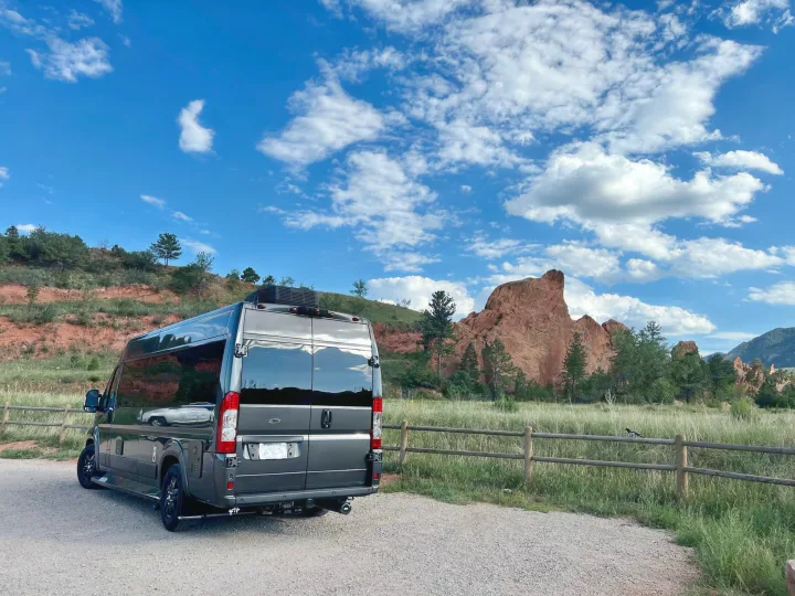 RV rental parked on gravel. A dark camper van with reflections overlooks red rock formations, green fields, & blue sky.. Dodge Other 2020