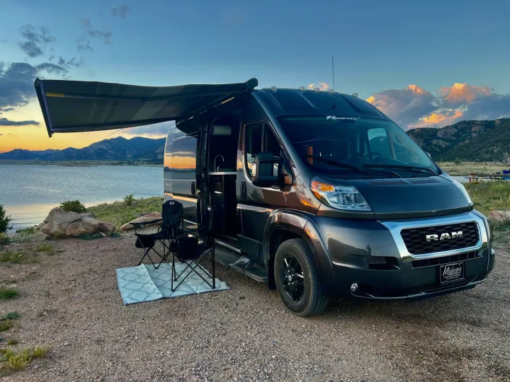 Stylish dark gray rv rental camper van parked by a scenic lake at sunset with mountains and camping chairs set up.. Dodge Other 2020