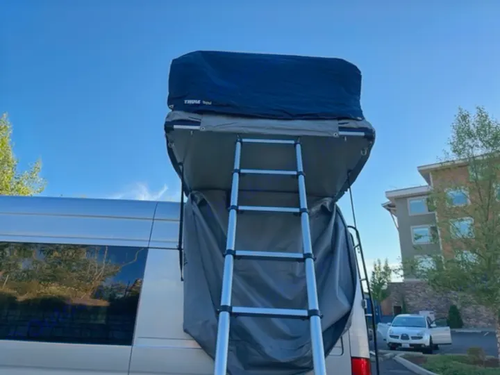 Back view of an RV rental van with a ladder leading to a deployed Thule rooftop tent for camping.. VanCraft Mercedes Benz 2022
