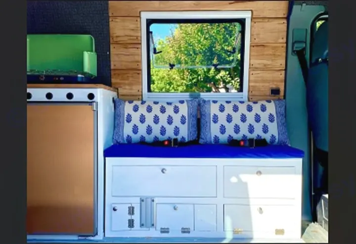 Cozy RV rental interior featuring a compact camper kitchen, comfortable blue seating with storage, and a scenic window. Perfect for van life.. Mercedes Sprinter 2005