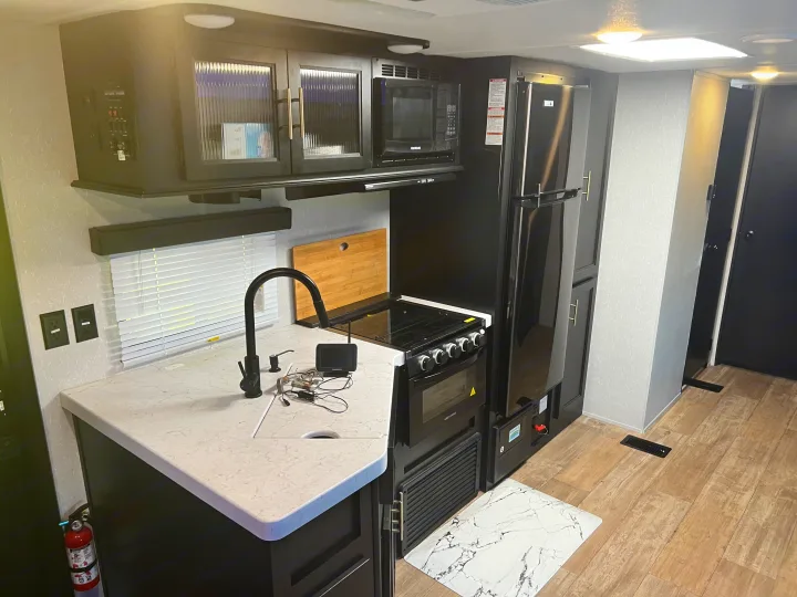 An RV rental's modern kitchen with black appliances, white countertops, and light wood floors. Includes a sink, stove, and fridge.. Forest River Cherokee 2021