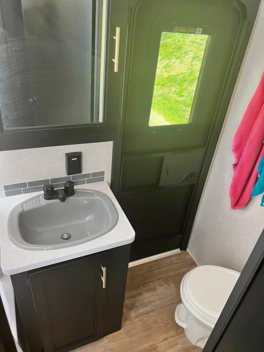 Compact RV rental bathroom features a gray sink, dark vanity, toilet, and an exterior door view.. Forest River Cherokee 2021