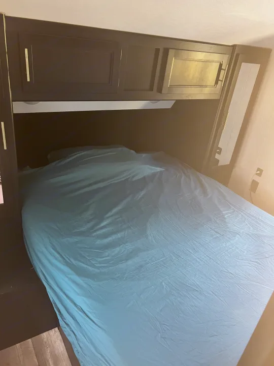 Inside an RV rental: a compact bedroom with a bed, teal sheet, and dark overhead cabinets.. Forest River Cherokee 2021