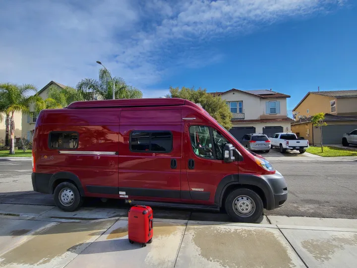 Red Winnebago Solis RV rental and a matching red suitcase on a sunny residential street.. Winnebago Solis 2022