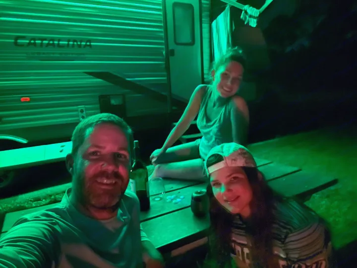 Friends enjoy a vibrant green-lit night outside their RV rental, next to a "Catalina" travel trailer and picnic table.. Forest River Coachmen Catalina Legacy 2022