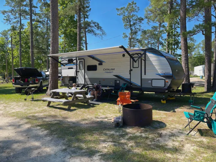 Family camping with an RV rental under pine trees. A picnic table, truck, and fire pit fill this sunny outdoor campsite.. Forest River Coachmen Catalina Legacy 2022