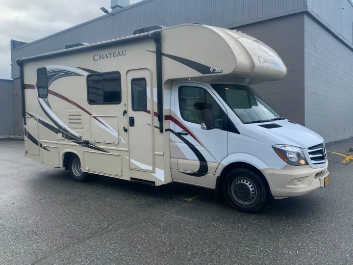 A Chateau Thor Class C RV rental, cream and white with stripes, is parked on asphalt beside a grey building.. Thor Motor Coach Chateau Sprinter 2018