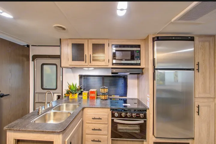 This modern RV rental kitchen features stainless steel appliances (fridge, microwave, stove), a dual sink, and light wood cabinetry.. Heartland Prowler 2022