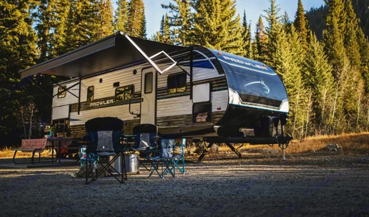 Ready for nature! This Prowler RV rental is perfectly set up at a sun-drenched forest campsite with chairs and a table.. Heartland Prowler 2022