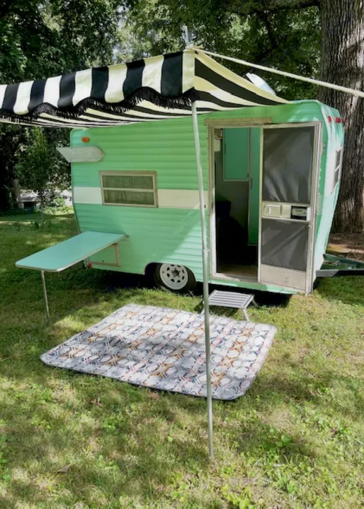 Retro green RV rental with striped awning & side table. Mat on grass for cozy outdoor escape.. Shasta Compact 1973