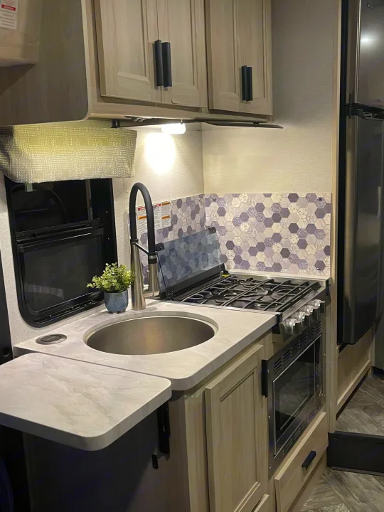 RV rental kitchen with a modern design: light wood cabinets, stainless sink, stove, oven, microwave, and purple hex tile backsplash.. Forest River Sunseeker LE 2022