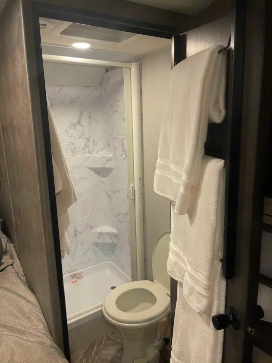 Compact RV rental bathroom: walk-in shower with marble walls, toilet, and fresh white towels hanging. Bed visible to the left.. Forest River Sunseeker LE 2022