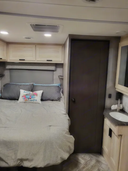 Cozy RV rental interior featuring a bed with overhead cabinets, a compact bathroom vanity with a sink, and a dark door.. Forest River Sunseeker LE 2022