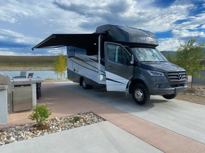 Grey Tiffin Wayfarer RV rental with extended awning parked lakeside. Features an outdoor kitchen for luxury camping.. Tiffin Motorhomes Wayfarer LW 2023