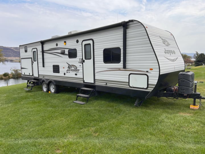 This Jayco travel trailer RV rental sits at a green lakeside campsite with mountains in the background.. Jayco Jay Flight 2016