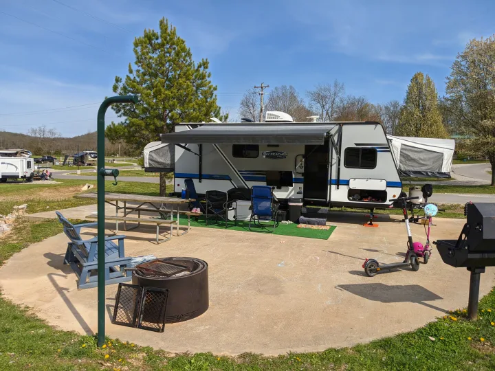 An RV rental travel trailer is set up at a sunny campsite with an outdoor rug, chairs, picnic table, fire pit, and grill.. Jayco Jay Feather 2019
