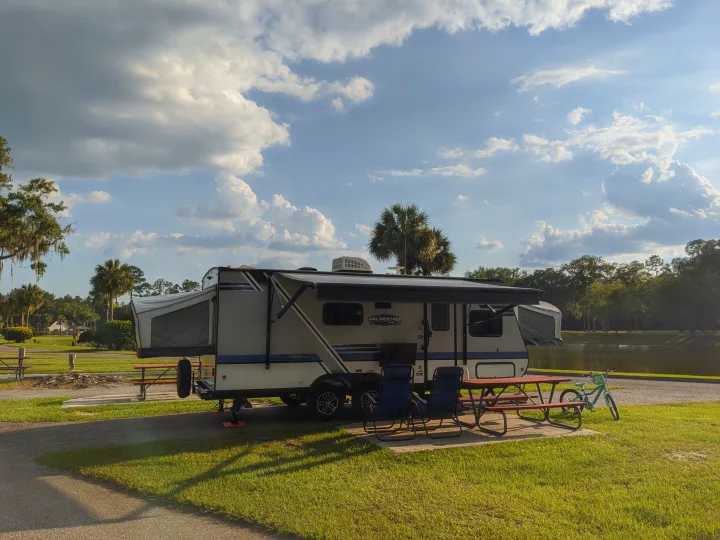 Relaxing RV rental campsite by a lake, featuring a hybrid travel trailer, picnic table, and bicycle under a sunny sky.. Jayco Jay Feather 2019