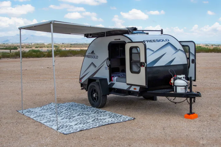 An RV rental: a grey teardrop trailer with an extended awning, patterned rug, and open doors, parked in a desert area.. Braxton Creek Free Solo 2022