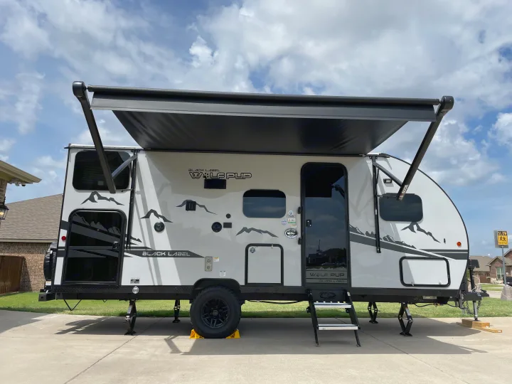 A white "Wolf Pup" travel trailer, available as an RV rental, set up with its extended awning and leveling jacks.. Forest River Cherokee Wolf Pup Black Label 2022
