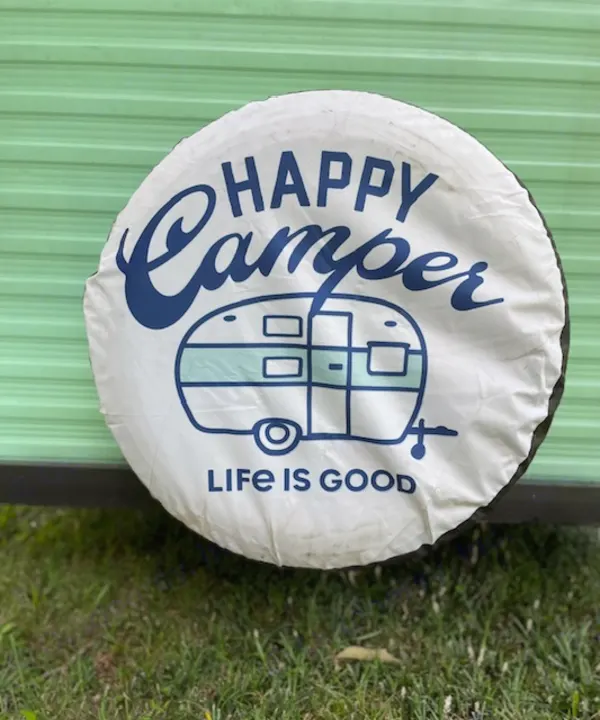 White spare tire cover with "Happy Camper, Life Is Good" text & a retro camper graphic on a green RV.. Shasta Compact 1973